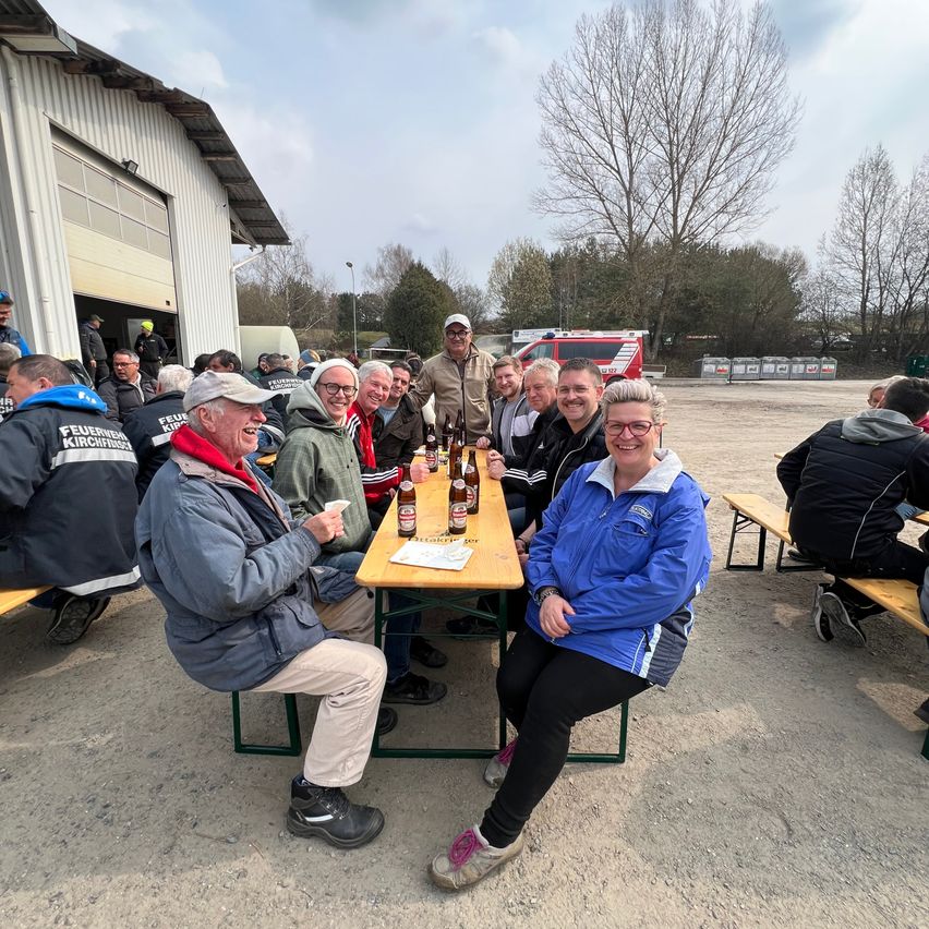 A group of people sit at a table outside, smiling. Bottles and a menu are on the table. Trees and a fire truck are in the background.