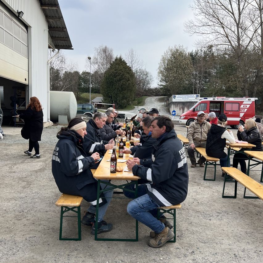 A group of people sit at outdoor tables, wearing jackets, some with reflective stripes. Bottles and food are on the tables, with a building and parked vehicles in the background.