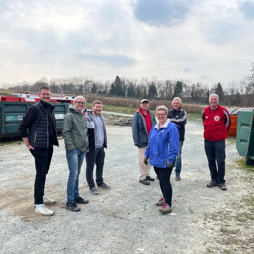 A group of six individuals stands in a gravel area, with three women and three men. They are dressed for cooler weather, and there are garbage bins in the background.