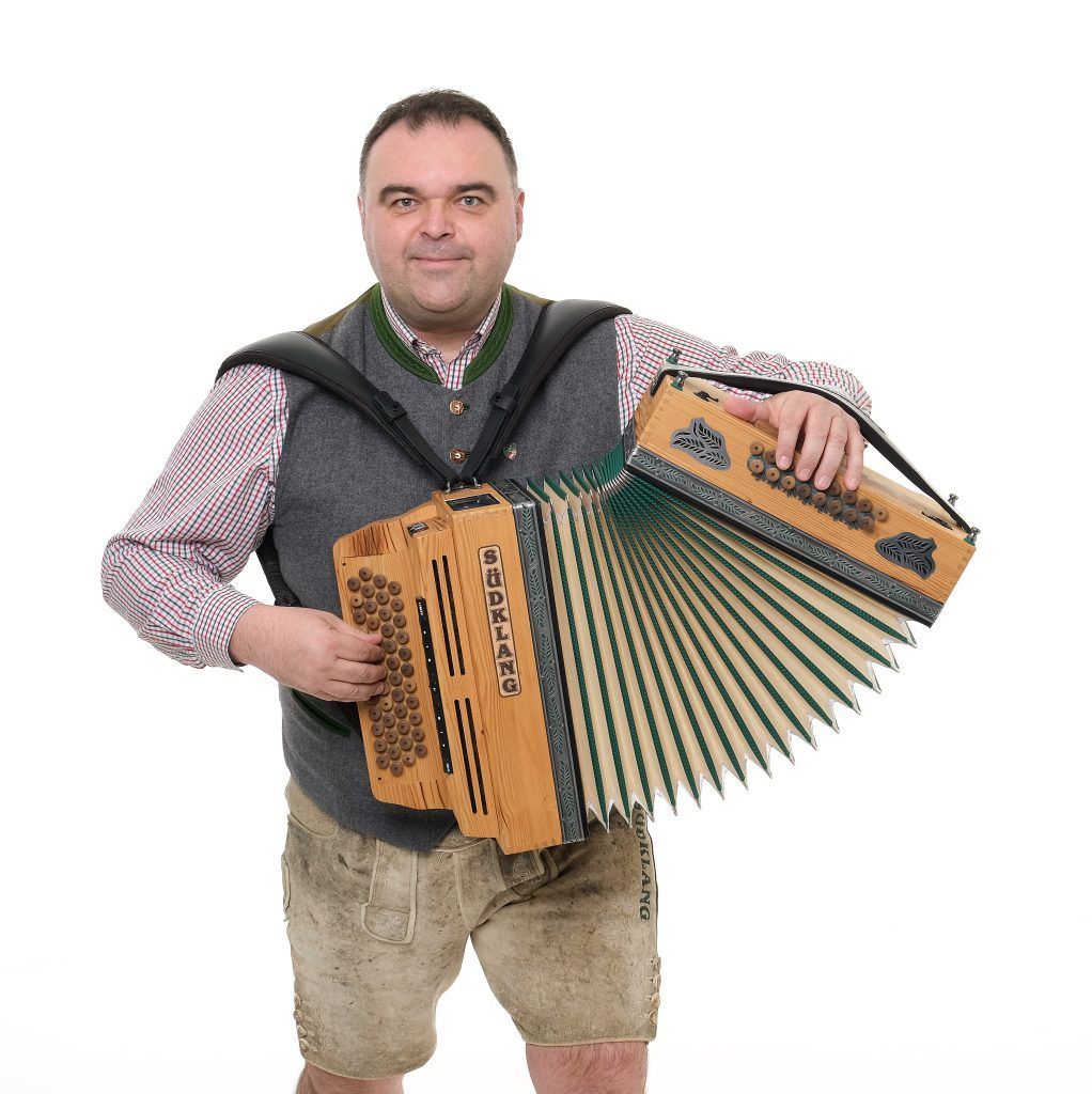 A man in a traditional Bavarian outfit is holding an accordion with the word Sudling written on it. He is smiling and posing for a photo.