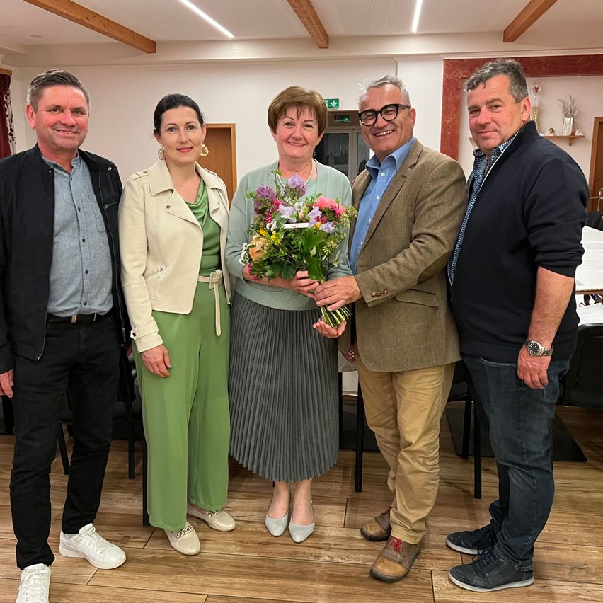 Five people stand indoors, smiling for a photo. The woman in the center holds flowers. Behind them are chairs and a table.