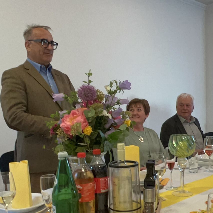 Three individuals, two men and a woman, are seated at a table. A man stands behind them, and there is a flower arrangement in the center. Bottles and wine glasses are also present.