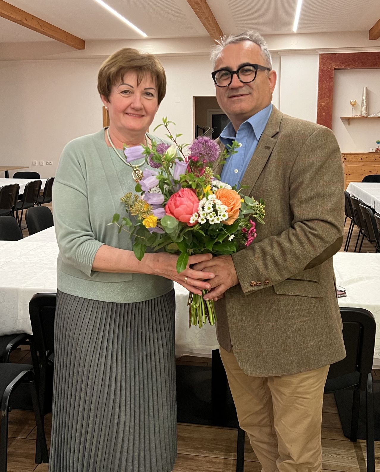 An adult man and woman holding a bouquet of flowers stand in a room with a table and chairs. The woman wears a necklace.