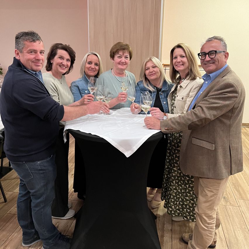 A group of smiling adults, both men and women, stand around a table holding wine glasses. They appear to be celebrating an event, possibly a meeting or gathering.