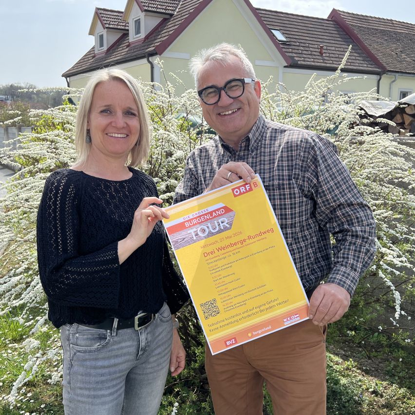 A man and a woman stand in front of a house, smiling and holding a yellow poster. The man wears glasses and a checkered shirt, while the woman wears a black blouse. The house has a brown roof and white walls.