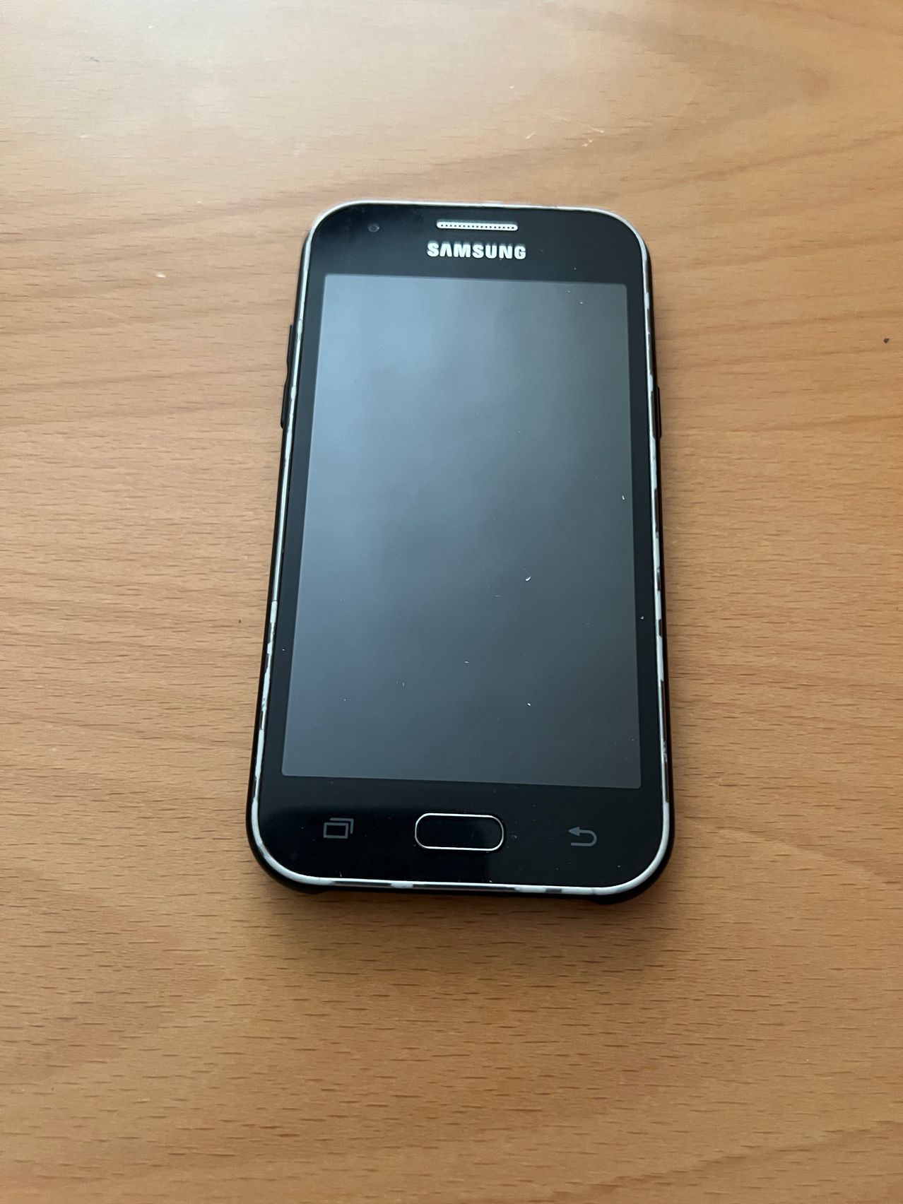 A Samsung smartphone, turned off, rests on a wooden surface. It has a home button and a power button.