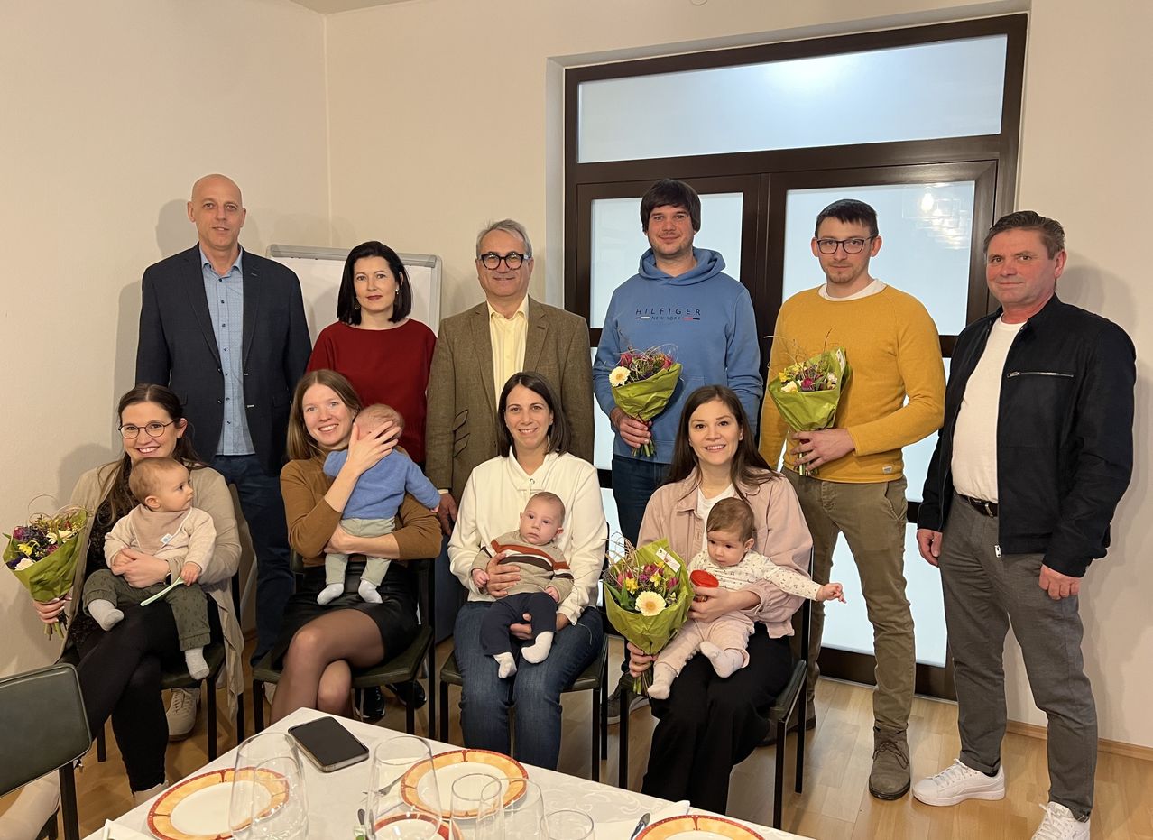 A group of people are gathered indoors, possibly for a family event. They are holding babies, and some are standing while others are sitting. There is a table with plates and glasses in front of them. The room has white walls and a glass door.