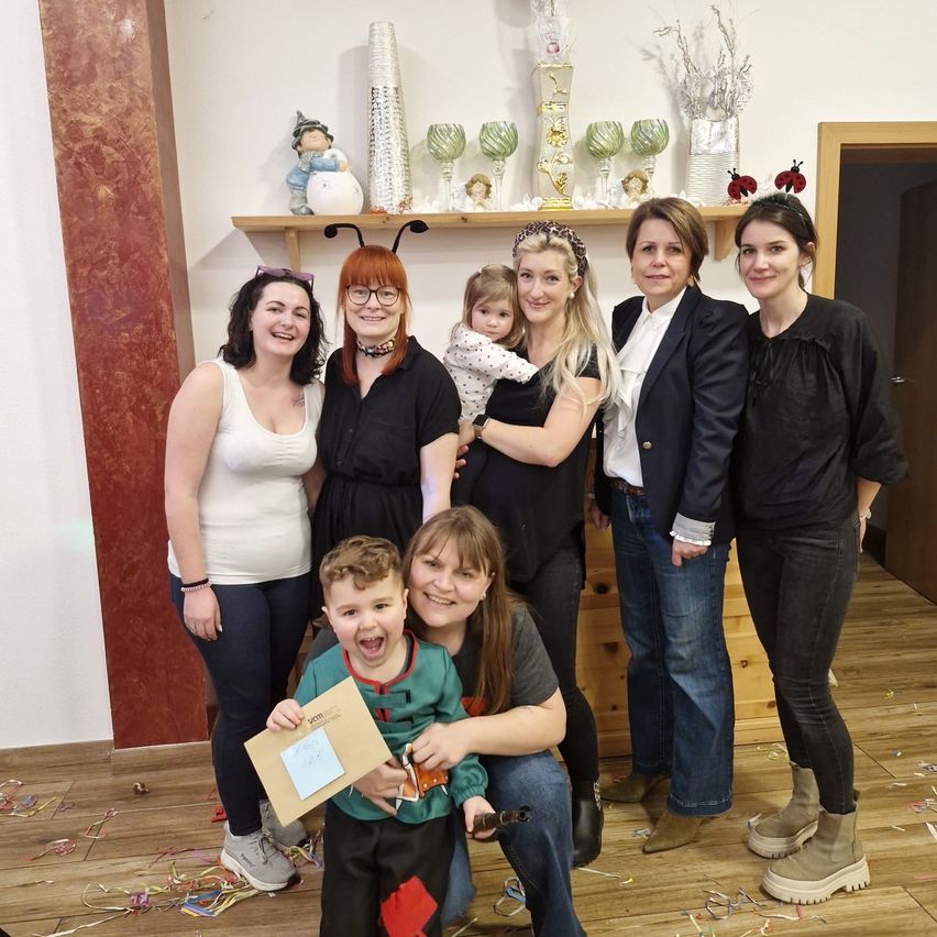 A group of women and children pose for a photo with a child holding a certificate, smiling and standing in a room with a wooden floor and a shelf with various items.