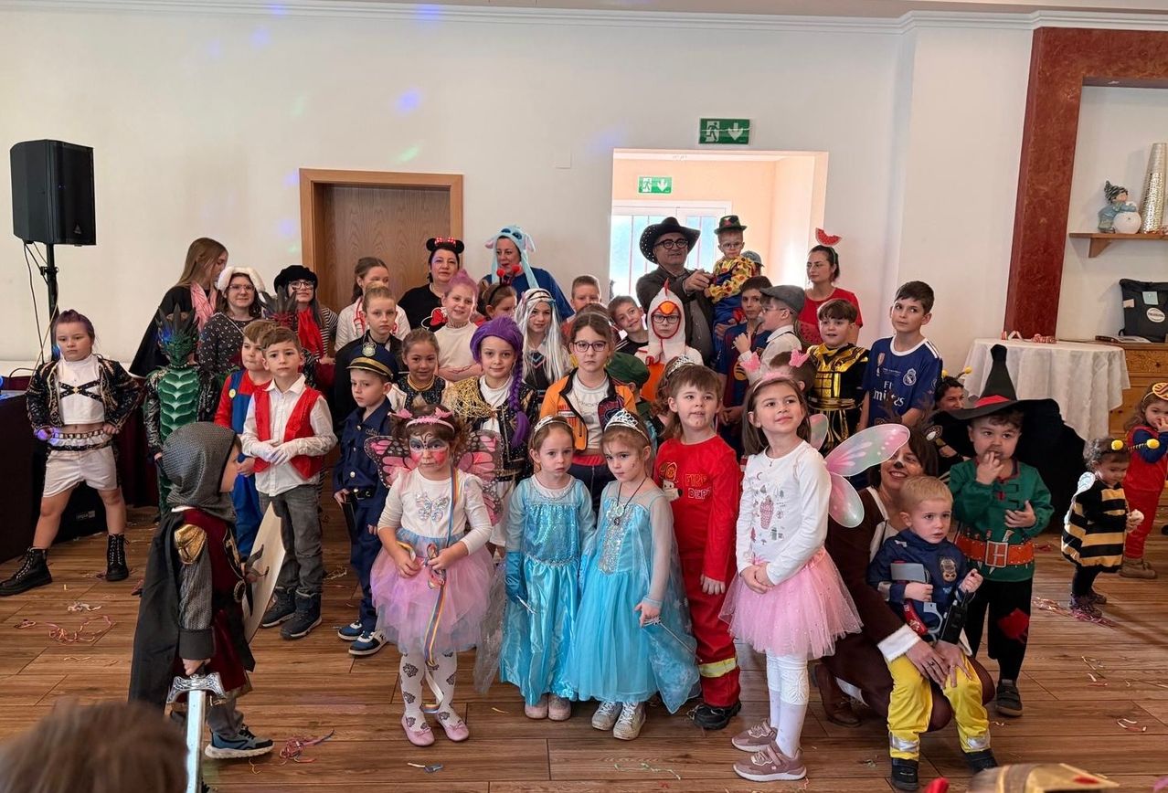 A large group of children in costumes, some holding props, are posing for a photo in a room with wooden floors and white walls. Some are dressed as fairies, while others are in superhero costumes. A man in a hat stands in the center.