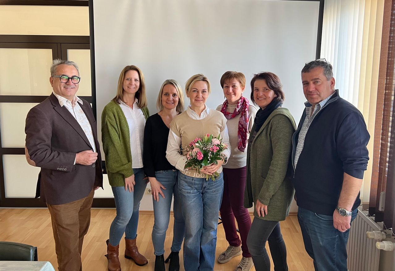 Eine Gruppe von sieben Menschen steht drinnen. Eine Frau in der Mitte hält einen Blumenstrauß. Alle lächeln und scheinen für ein Foto zu posieren.