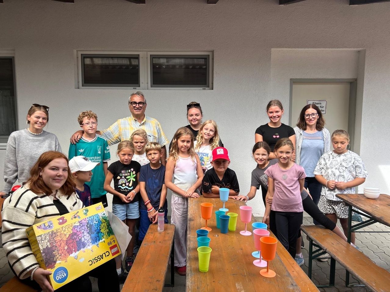 A group of children and adults are gathered around a wooden table with colorful cups, posing for a photo. The children are smiling and some are holding a box. The adults are standing behind them. The wall behind them has a window with blinds.