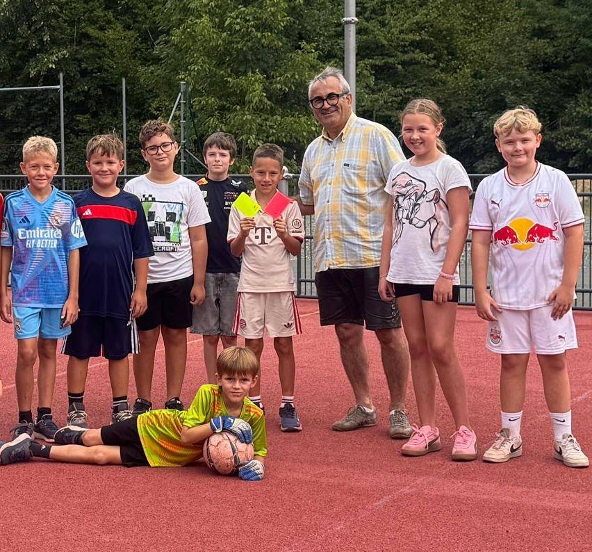 A group of children and an adult male are standing on a red field, likely a soccer field. The children are wearing different colored jerseys, and one of them is holding a soccer ball. The adult male is wearing glasses and a checkered polo shirt.