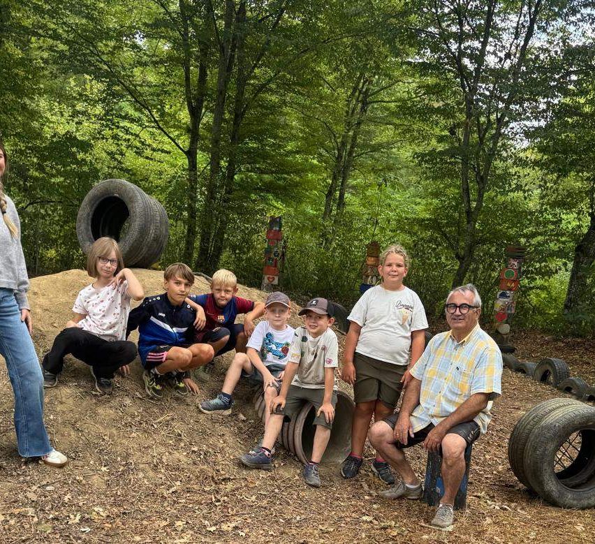 A group of children and an adult man are posing for a photo in a forest setting with large tires and wooden posts.
