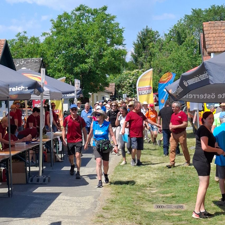 Eine Menschenmenge, einige mit Hüten und Sonnenbrillen, geht an einem sonnigen Tag an einem Event mit Zelten und Tischen vorbei.
