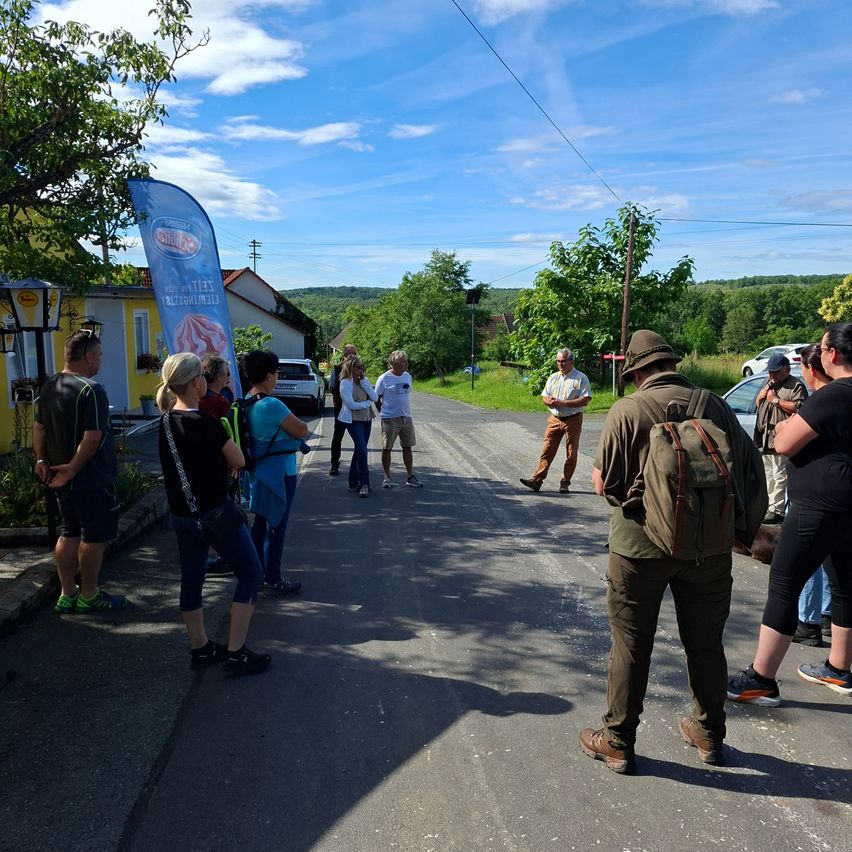 Eine Gruppe von Menschen steht auf einer Straße, einige blicken auf ein Banner. Autos sind in der Nähe geparkt. Die Szene spielt sich an einem sonnigen Tag mit klarem Himmel ab.