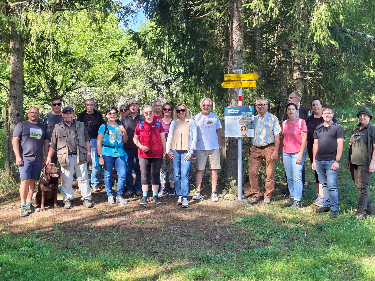 Eine Gruppe von Menschen steht in einem Wald und lächelt für ein Foto. Ein Schild befindet sich hinter ihnen.