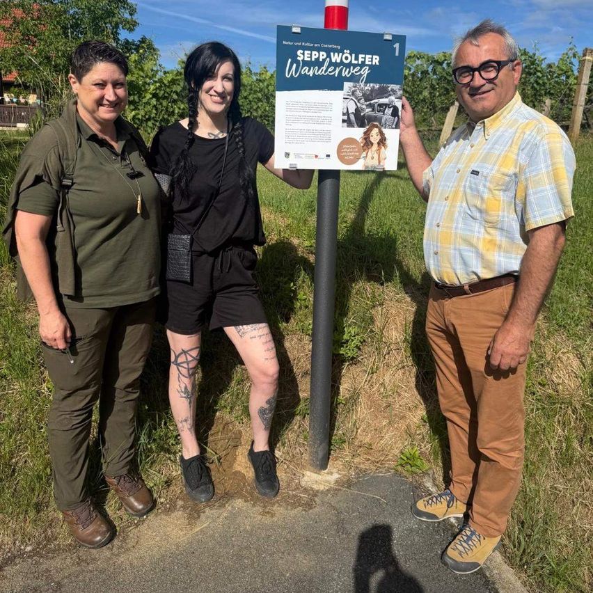 Drei Menschen stehen bei einem Schild, zwei Frauen und ein Mann, lächelnd für ein Foto auf einem Grasfeld mit einem Weinberg im Hintergrund.