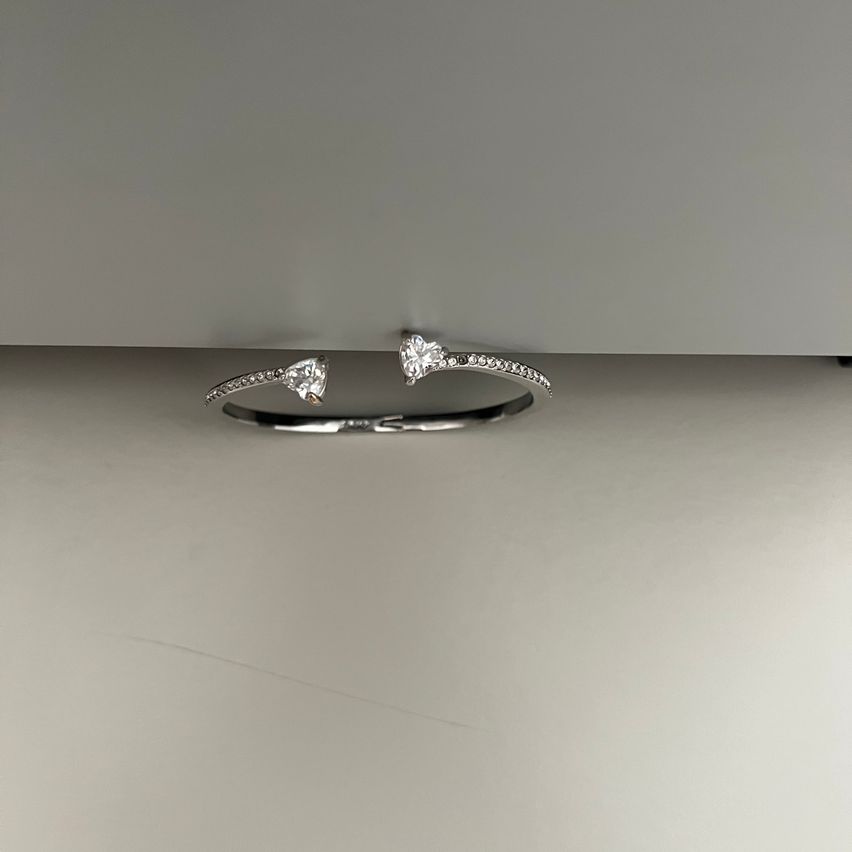 Ein silbernes Armband mit zwei herzförmigen Diamantakzenten, das auf einer grauen Oberfläche ruht. Das Armband ist gebogen und hat eine leicht glänzende Erscheinung.