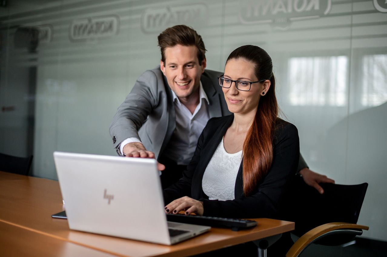 A man and a woman are sitting at a desk, the man is leaning over the woman who is working on a laptop.