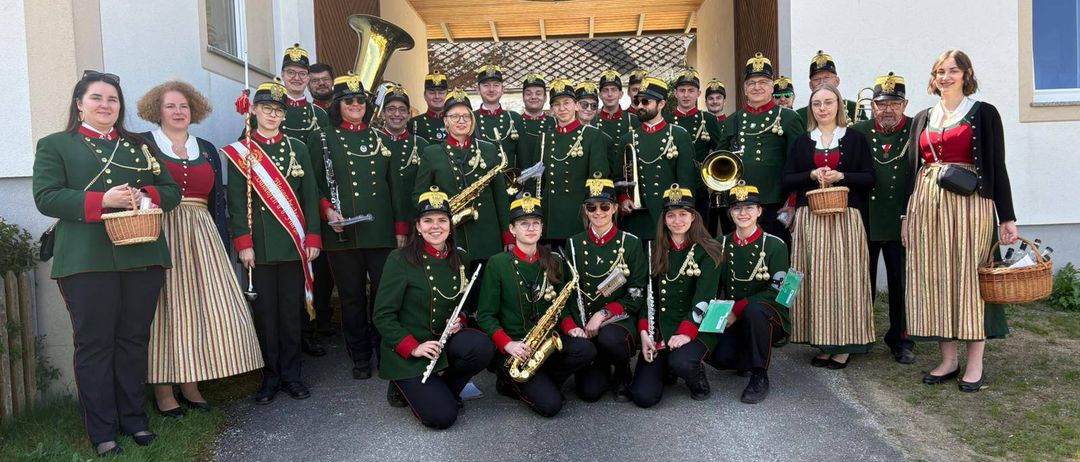 Ein Blasorchester und eine Brass Band posieren in traditionellen grünen Uniformen für ein Foto, einige kniend, andere stehend, mit Musikinstrumenten. Der Hintergrund zeigt ein Gebäude, eine Lampe und Menschen.