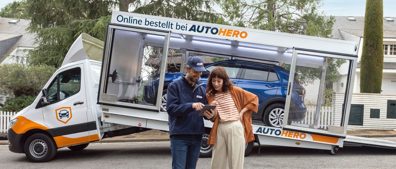 Ein Mann und eine Frau stehen neben einem Autotransporter und schauen auf ein Tablet. Der Anhänger ist mit 'Online bestellt bei AUTOHERO' beschriftet.