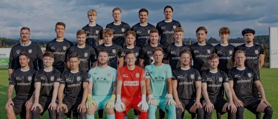 Eine Fußballmannschaft posiert für ein Gruppenfoto auf einem Rasenfeld, wobei die Spieler schwarze und blaue Trikots tragen. Der Torwart ist in einem roten Trikot.