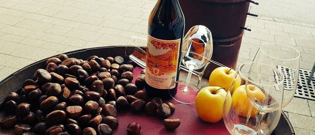 Eine Flasche Wein mit rotem Etikett steht neben einem roten Tischset, umgeben von Kastanien, Äpfeln und Weingläsern. Im Hintergrund befindet sich ein Fass.
