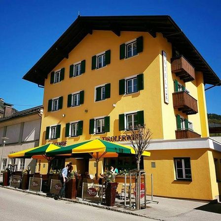 Ein gelbes Gebäude mit grünen Fensterläden, beschriftet mit Hotel Tirolerwirt, steht in einer Stadt. Es hat einen Balkon und eine Terrasse mit sitzenden Menschen. Der Himmel ist blau.