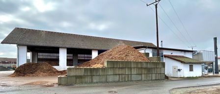 Ein Bauernhof mit einem Haufen Holzspäne und einer Betonbarriere davor, unter einem bewölkten Himmel.
