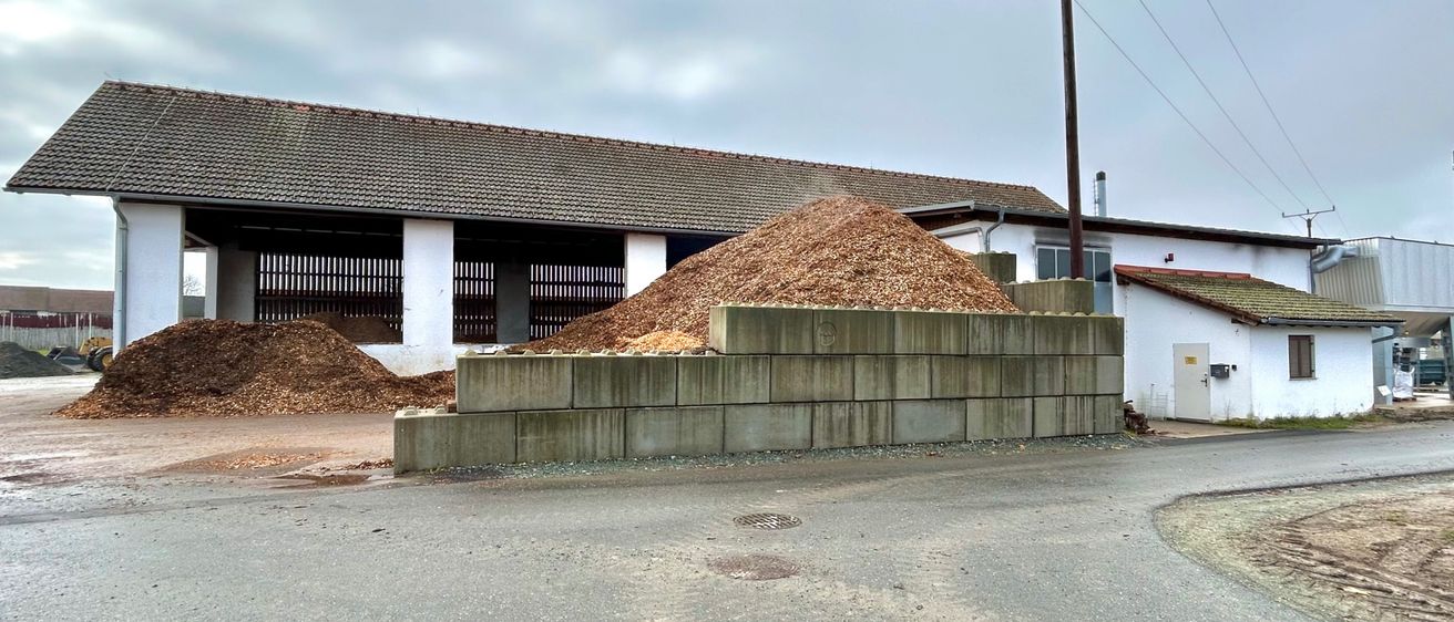 Eine ländliche Szene mit einem großen Haufen von Holzspänen vor einer Scheune unter einem bewölkten Himmel mit Stromleitungen im Hintergrund.