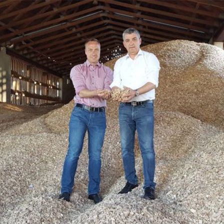 Zwei Männer stehen in einer Scheune und halten eine Ansammlung von Holzspänen. Einer hält eine Handvoll, während der andere die Qualität zeigt.