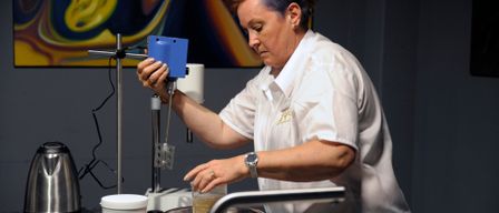 Eine Frau mit blauen Haaren arbeitet in einem Labor. Sie hält ein blaues Gerät und gießt Flüssigkeit in eine Schüssel. Sie trägt ein weißes Hemd und hat Flaschen und Werkzeuge auf dem Tisch.