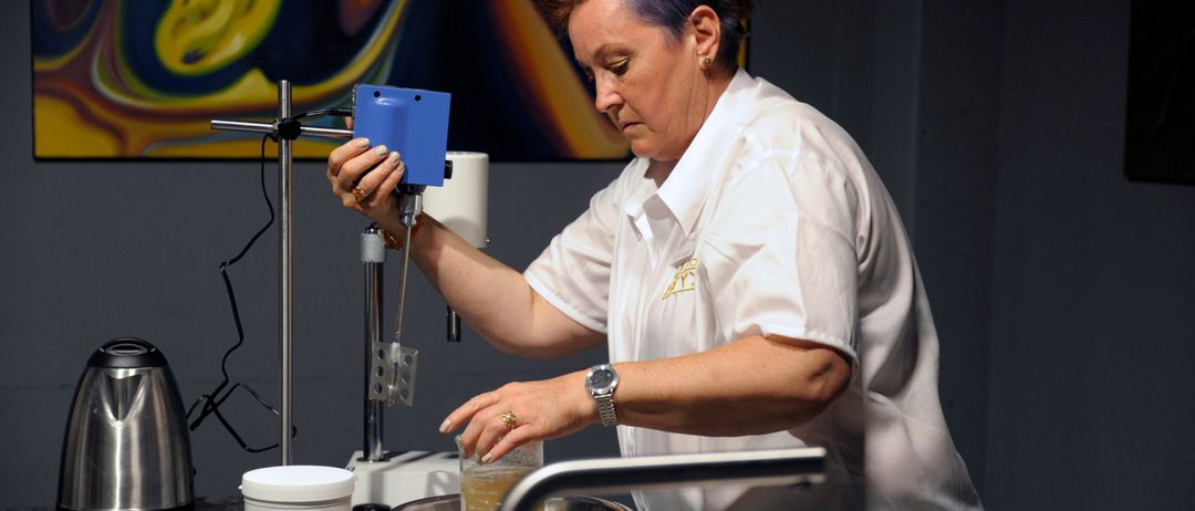 Eine Frau mit blauen Haaren arbeitet in einem Labor. Sie hält ein blaues Gerät und gießt Flüssigkeit in eine Schüssel. Sie trägt ein weißes Hemd und hat Flaschen und Werkzeuge auf dem Tisch.