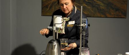Eine Frau mit kurzen Haaren arbeitet an einem wissenschaftlichen Experiment. Sie benutzt eine Maschine, um Flüssigkeiten in einem Becher und einem Kolben zu mischen.