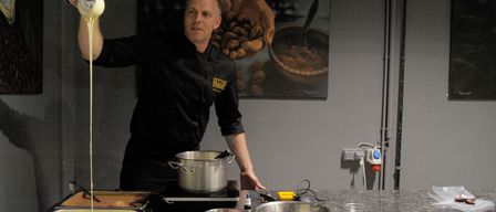 Ein Koch gießt eine Flüssigkeit in einen Topf. Mehrere Schüsseln und ein Tablett stehen auf der Arbeitsplatte. An der Wand befinden sich eine Steckdose und eine Rohrleitung.