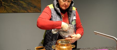 Eine Frau in einem roten Shirt und einer grauen Weste gießt Zutaten in einen Kupfertopf auf dem Herd. Der Topf hat einen Deckel und steht auf einem Brenner. Ein Kupfertopf mit Deckel steht links auf dem Herd.