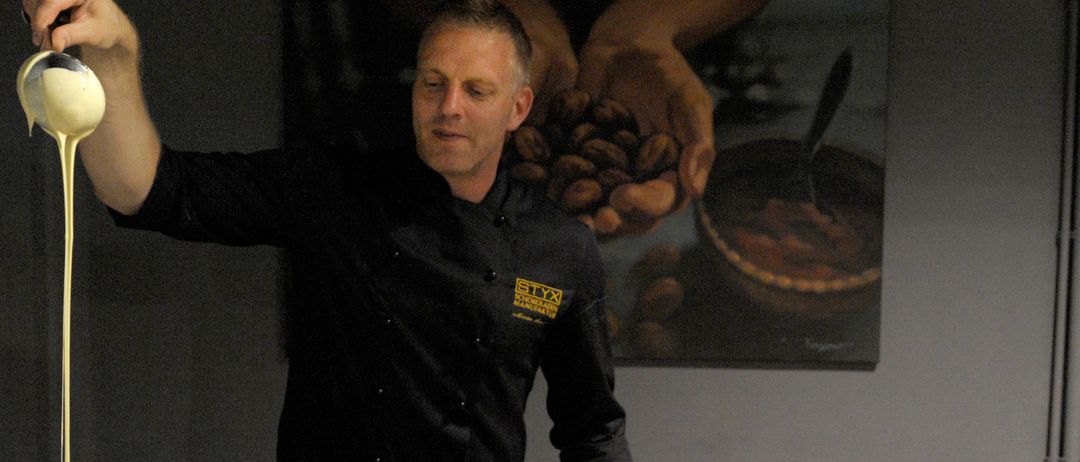 Ein Koch in einer schwarzen Uniform steht an einem Küchentresen mit Kochutensilien und Zutaten und bereitet Essen in einem Topf zu.