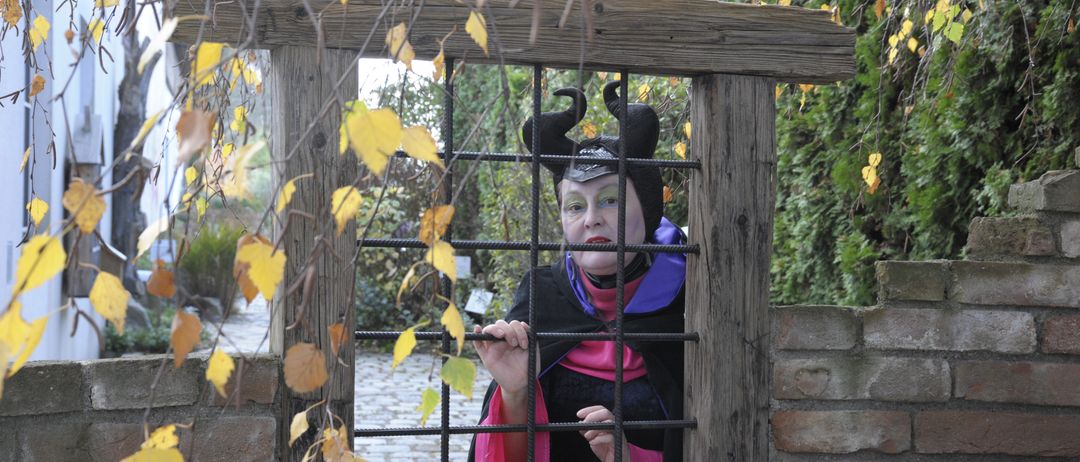 Eine Person, die als Malefiz verkleidet ist, steht hinter einem Holztor und hält die Gitterstäbe mit beiden Händen fest. Der Ort ist draußen mit Herbstblättern und einer Ziegelmauer.