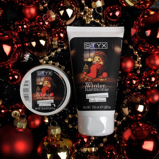 Eine Nahaufnahme einer Winter Harmonia Körpercreme und eines kleinen runden Behälters vor einem Hintergrund roter Ornamente.