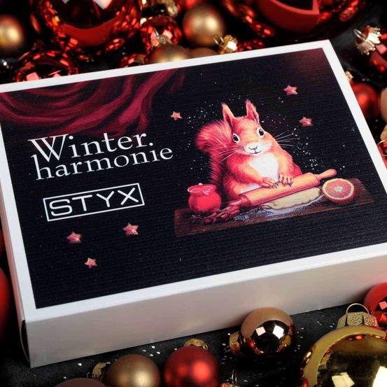 Eine weiße Box mit dem Markennamen STYX. Die Box enthält ein Winterharmonie-Set. Die Box hat ein Bild von einem Eichhörnchen, das einen Nudelholz hält und eine Orange. Der Hintergrund ist dunkel und es gibt Sterne und Ornamente.