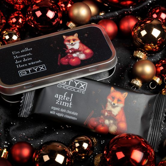 Eine silberne Blechdose mit einer Fuchsillustration, die Schokoladenriegeln enthält. Der Schokoladenriegel hat Text, der 'Apfel Zimt' und 'Bio-dunklen Schokolade mit Apfel-Zimt' lautet. Es gibt Weihnachtskugeln drumherum.