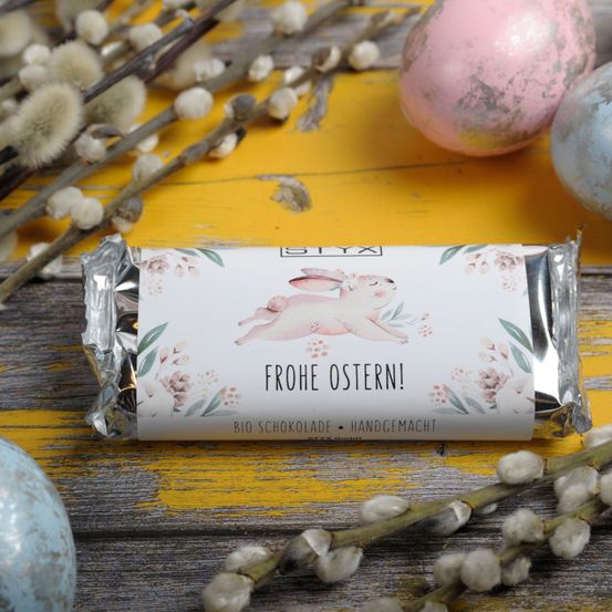 Eine in Alufolie verpackte Süßigkeit mit Ostereiern und Weidenzweigen auf gelbem Hintergrund. Es steht Frohe Ostern! auf Deutsch.