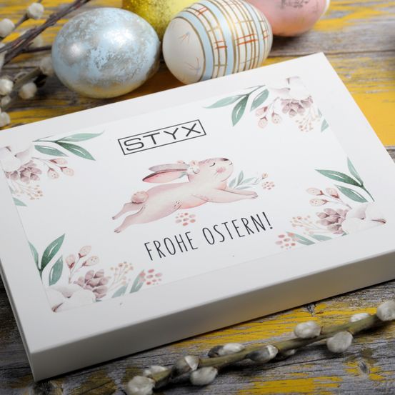 Eine weiße Box mit Blumen- und Hasendesign sagt 'Frohe Ostern!' darauf. Hinter ihr befinden sich Ostereier.