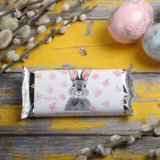 Eine vegane Schokoladentafel mit einem Hasenbild liegt auf einem gelben Holztisch mit Ostereiern und Weidenzweigen.