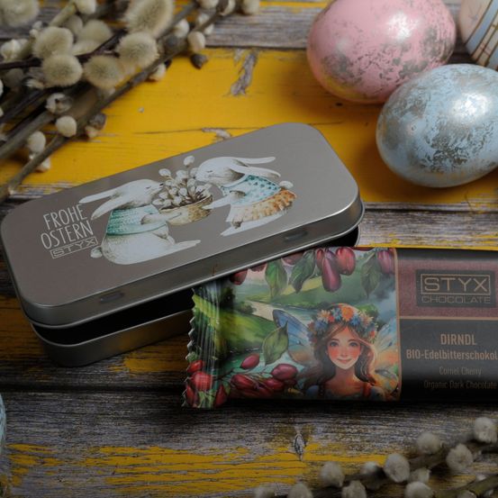 Eine Blechdose mit einer Hasenillustration, die eine Schokoladentafel mit einer Fee enthält. Verschiedene Ostereier liegen auf einer gelben Oberfläche.