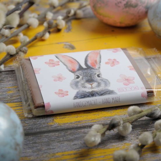 Eine Schokoladentafel mit einem Hasen auf der Verpackung liegt auf einer gelben Oberfläche. Sie ist von Ostereiern und Zweigen mit weißen Blüten umgeben. Die Schokoladentafel befindet sich in Plastikverpackung.