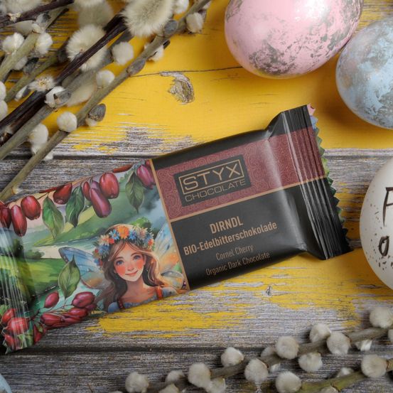 Eine Schokoladentafel mit einem Elfenmädchen auf dem Cover liegt auf einem gelben Tisch neben einigen Ostereiern. Die Schokolade ist bio und hat einen Kornelkirschengeschmack.