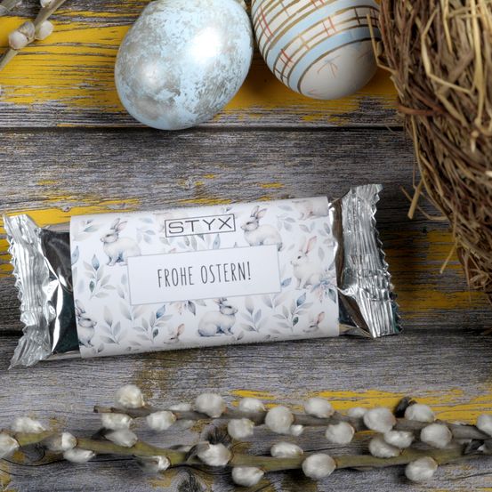 Eine silberumwickelte STYX-Schokoladentafel mit einem Osterhasen und Blumendesign liegt auf einer Holzoberfläche neben zwei Ostereiern und einem Nest.
