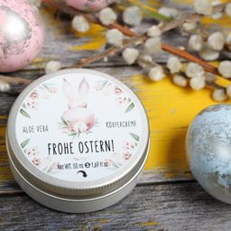 Eine Dose Körpercreme mit einem floralen Hasendesign auf dem Deckel, umgeben von Ostereiern und Frühlingsblumen auf einer Holzoberfläche. Das Etikett trägt die Aufschrift 'Aloe Vera Frohe Ostern!' und 'Nettogewicht 50 ml e 1,69 fl oz.'