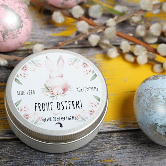 Eine Dose Körpercreme mit einem floralen Hasendesign auf dem Deckel, umgeben von Ostereiern und Frühlingsblumen auf einer Holzoberfläche. Das Etikett trägt die Aufschrift 'Aloe Vera Frohe Ostern!' und 'Nettogewicht 50 ml e 1,69 fl oz.'